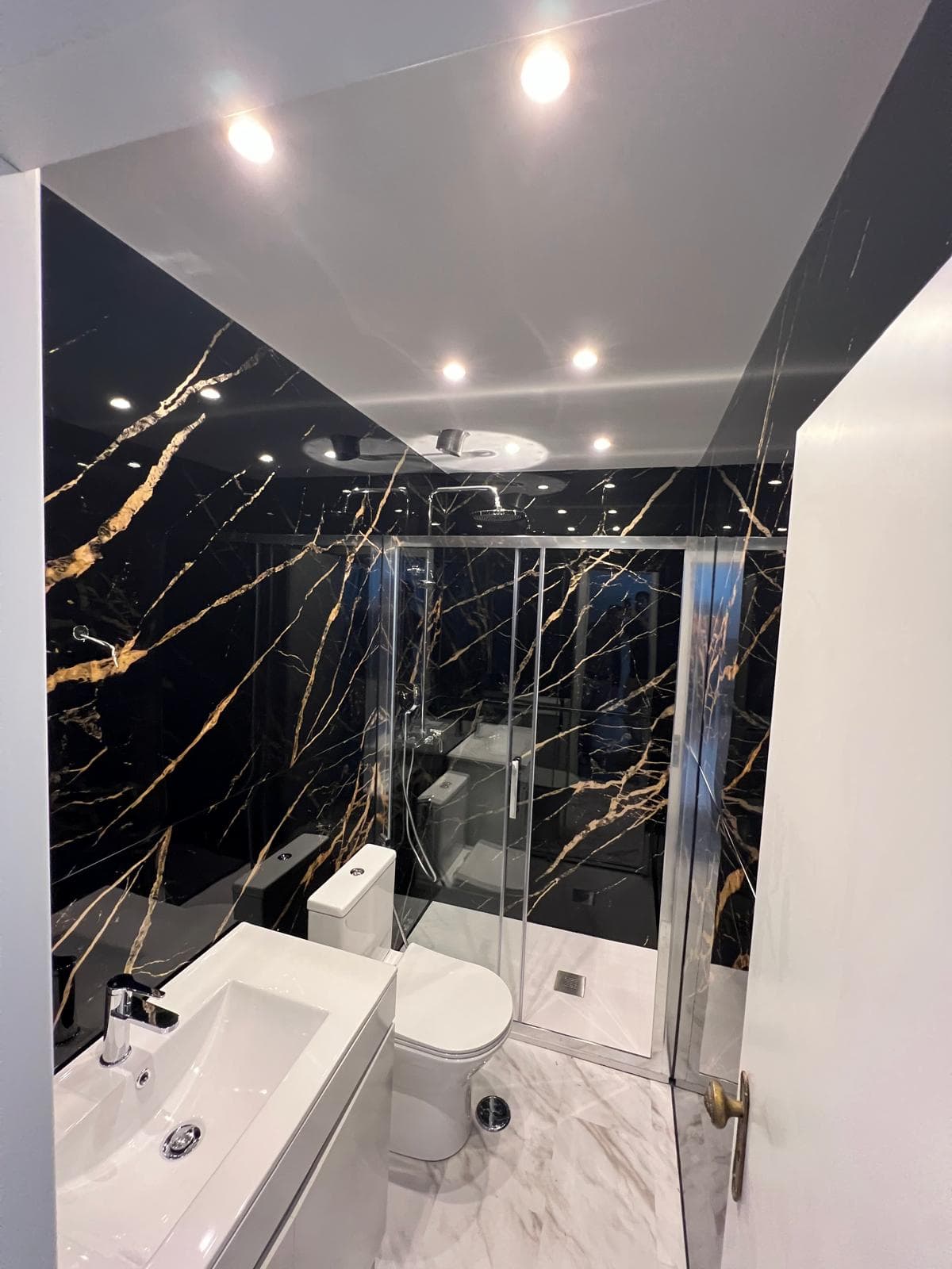 Casa de banho remodelada pela Elite Reparações em Lisboa, com revestimento em efeito mármore negro com veios dourados, duche com resguardo de vidro e iluminação encastrada no teto.